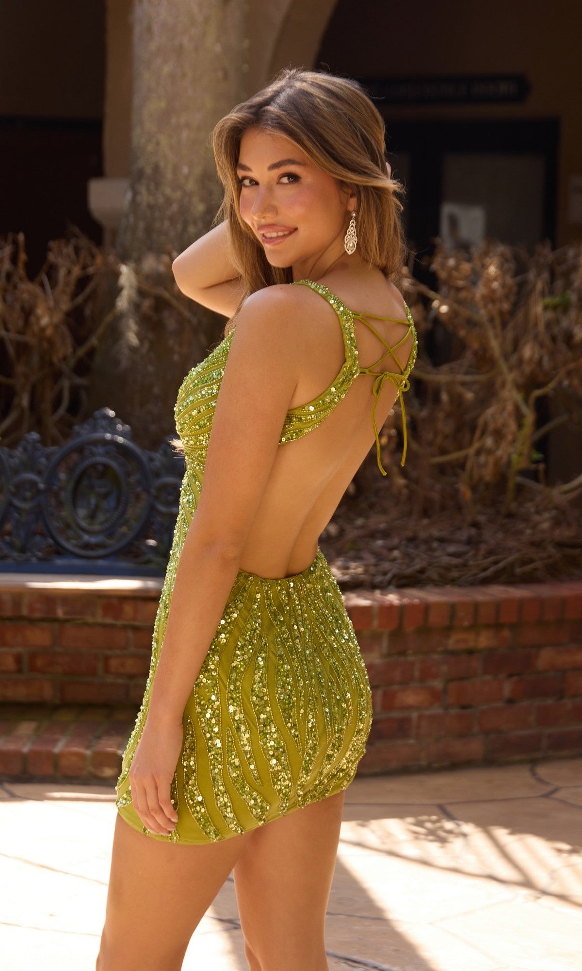 A woman in a backless, green Primavera 4694 short homecoming dress stands outdoors, looking over her shoulder and smiling.
